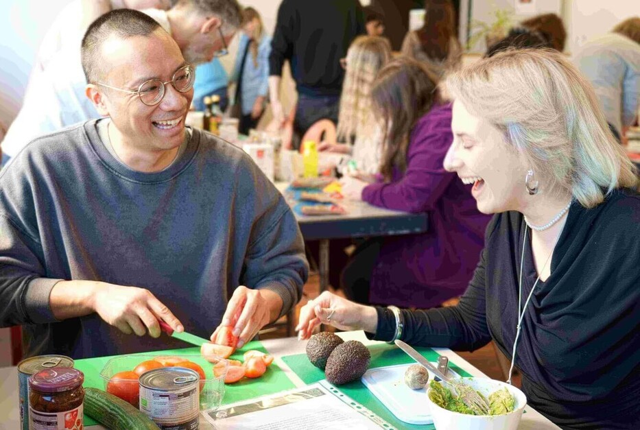Zwei Teilnehmende im Gespräch während der Essensvorbereitung, Gruppenaktivität im Hintergrund.